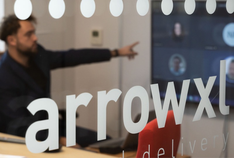 An ArrowXL colleague pointing at a TV meeting with the front of the image showing the ArrowXL logo etched on the meeting room glass